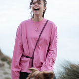 Lachende Frau im pinken Longsleeve „Rather be with“ mit Oversized Fit und Schriftzug auf dem Ärmel, aus 100 % Baumwolle und gefertigt in Portugal – dazu eine weinrote Umhängeleine.