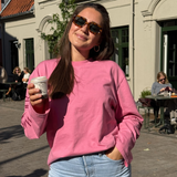 Lächelnde Frau mit Sonnenbrille und Kaffeebecher trägt das pinke Longsleeve „Rather be with“ im Oversized Fit mit Hej Nika Logo mittig auf der Brust, aus 100 % Baumwolle und gefertigt in Portugal – kombiniert mit heller Jeans.