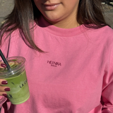 Detailaufnahme des pinken Longsleeves „Rather be with“ im Oversized Fit mit dunklem Hej Nika Logo auf der Brust, aus 100 % Baumwolle und gefertigt in Portugal – in der Hand ein Matcha-Drink.