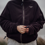 Frau trägt die Fleecejacke VARM in Chocolate Braun mit durchgehendem Reißverschluss, Stehkragen und gesticktem Hej Nika Logo auf der Brust – aus 100 % Baumwolle, gefertigt in Portugal. Gummizipper zum enger stellen von Ärmeln und Bund. 