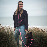 Frau in der Fleecejacke VARM in Chocolate Braun steht mit ihrem braunen Hund an pinker Leine in den Dünen am Meer – Die Jacke aus 100 % Baumwolle, gefertigt in Portugal.