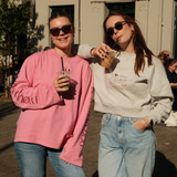 Frau trägt das pinke Oversized-Longsleeve „Rather be with“ aus 100 % Baumwolle mit Schriftzug auf dem Ärmel, kombiniert mit Jeans und Sonnenbrille – neben ihr eine Freundin im grauen Sweatshirt. Coole Looks für Hundebesitzer für Gassi oder auch zuhause. Oversized fit. 