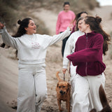 Frauen am Strand im Fokus eine Frau im Hellgrauen Jogger-Set von Hej Nika. Aus Baumwolle und recyceltem Polyester, hergestellt in Portugal. Hej Nika Print und Logo in Weinrot auf der Vorderseite.
