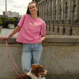 Frau trägt das pinke Hej Nika Dog Girls T-Shirt zu Jeans und steht mit einem braun-weißen Hund an der Spree in Berlin