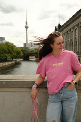 Frau im locker geschnittenen pinken Dog Girls T-Shirt mit Jeans und Hundeleine vor einer Berliner Stadtkulisse