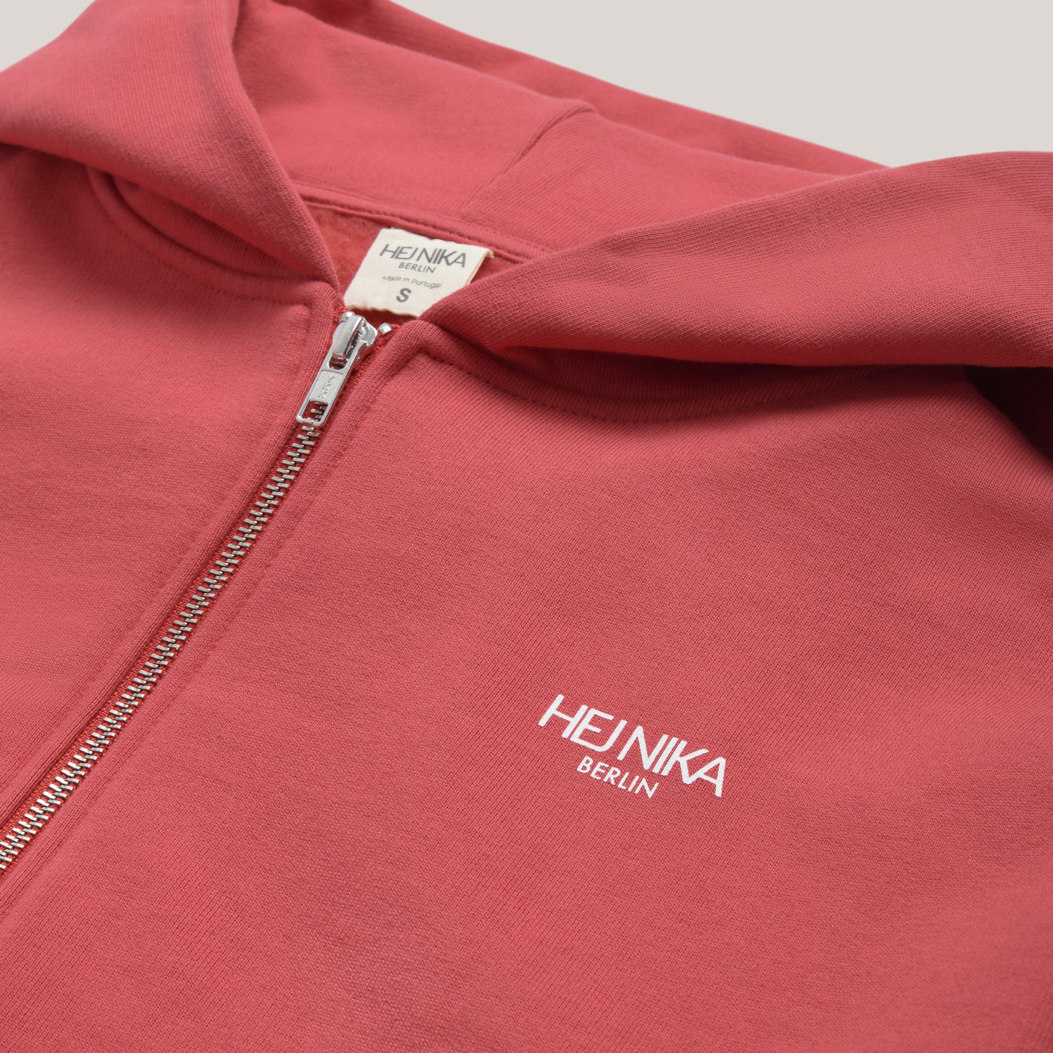 Detailansicht des silbernen Reißverschlusses und des weißen Hej Nika Logos auf der linken Brust der roten Sweatjacke.